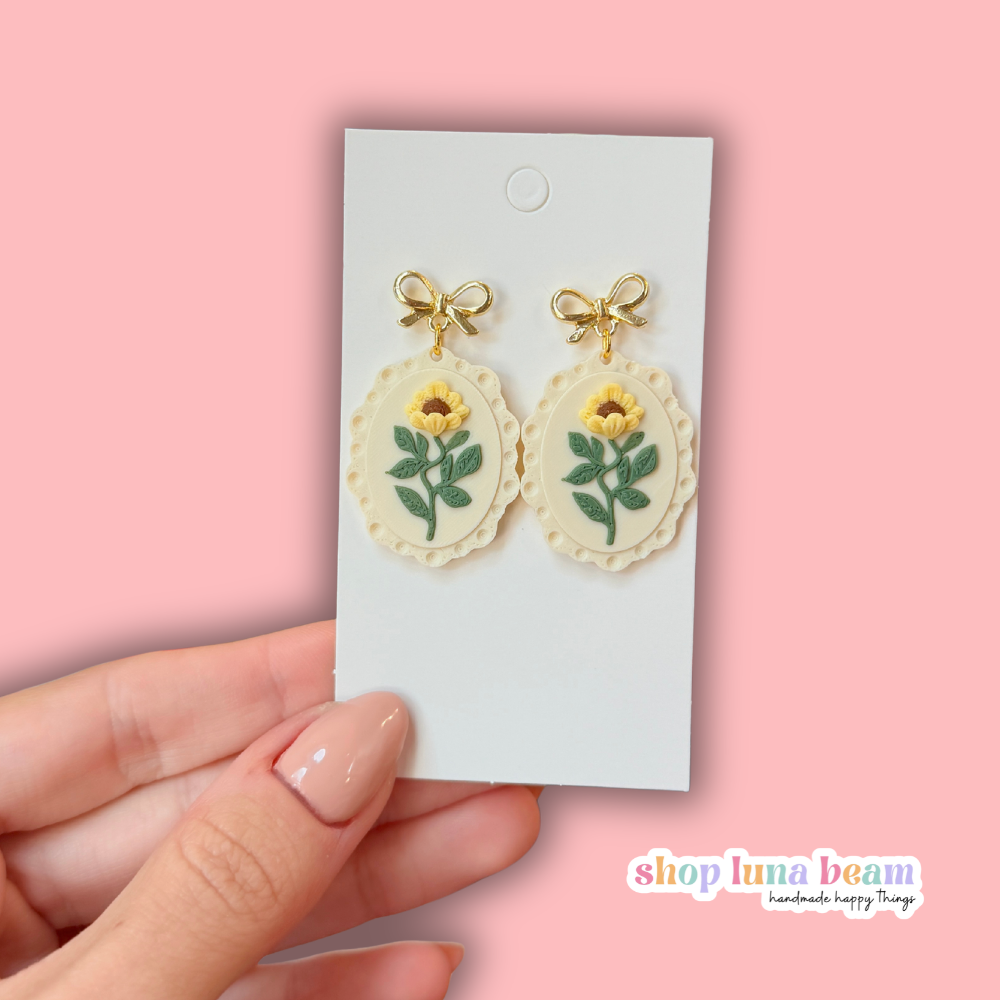 A pair of dangling earrings displayed on a white card, held by a hand against a soft pink background. Each earring has a small gold bow at the top and an oval cream-colored charm featuring a yellow flower with green leaves.