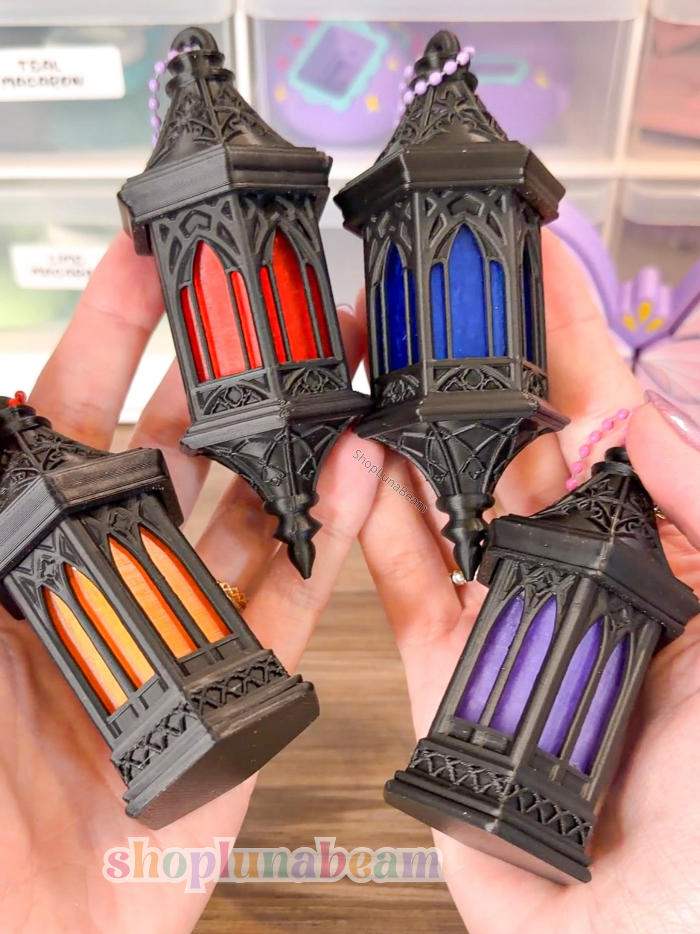 Four black lantern-shaped fidget clickers with colored glass held by hands against a blurred background.