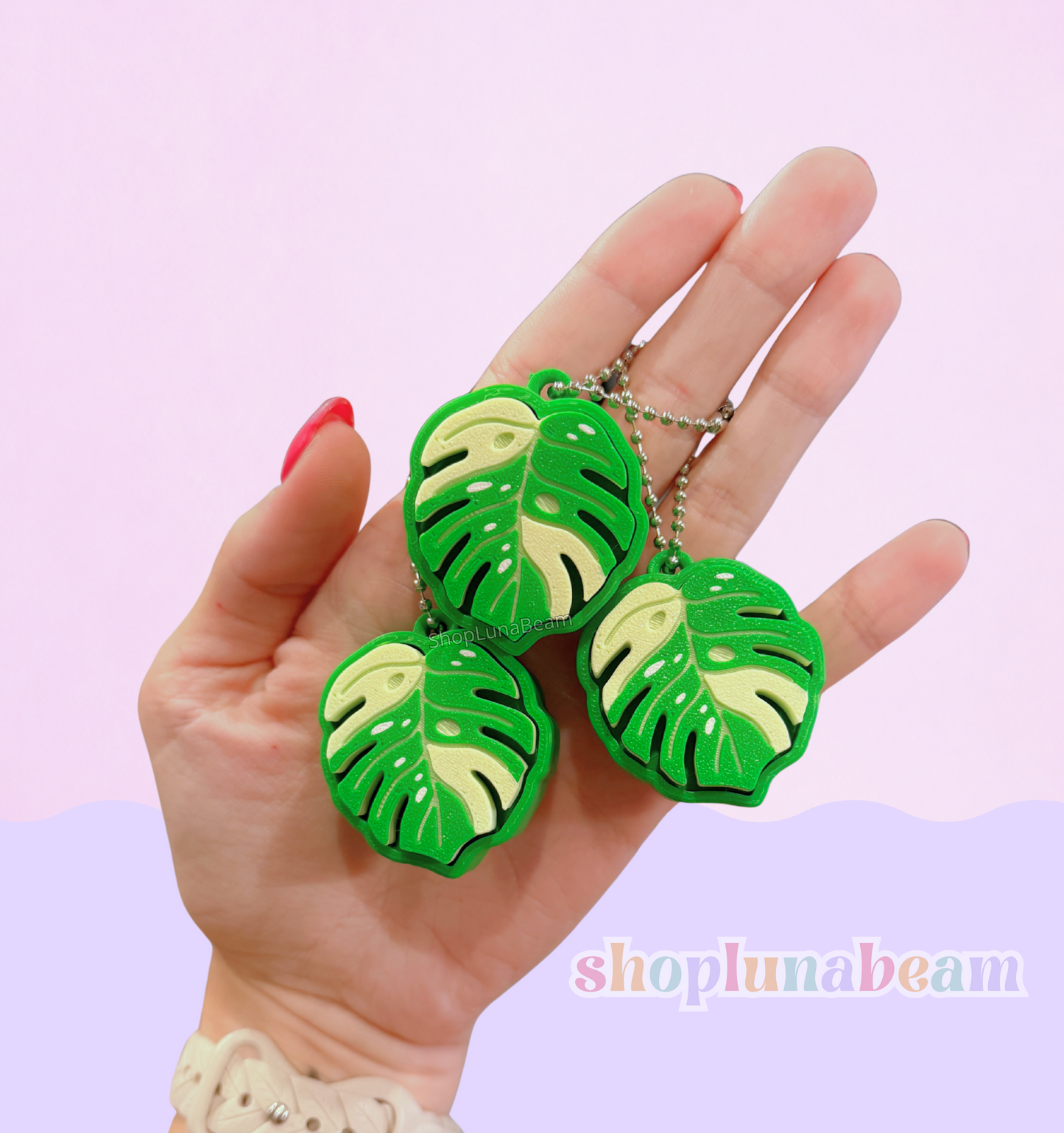 Hand holding three green leaf-shaped keychains against a light pink background with 'shopplumabeam' branding.