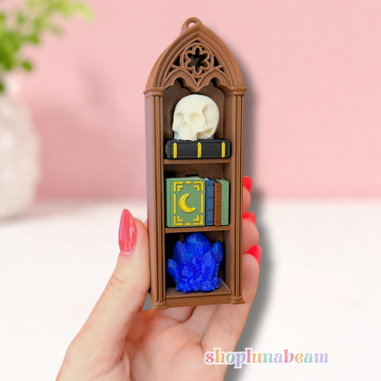 A hand with pink nail polish holds a miniature brown gothic bookshelf charm. The shelf contains tiny accessories including a white skull, a black book with gold trim, a green and gold book with a crescent moon, and a bright blue crystal, set against a soft pink and white background.