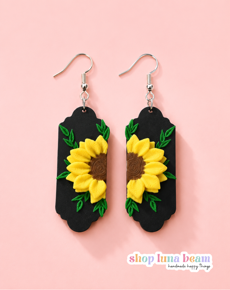 A pair of handmade dangle earrings featuring yellow sunflowers with brown centers and green leaves on a black background, suspended from silver fishhook wires, set against a solid pink background.