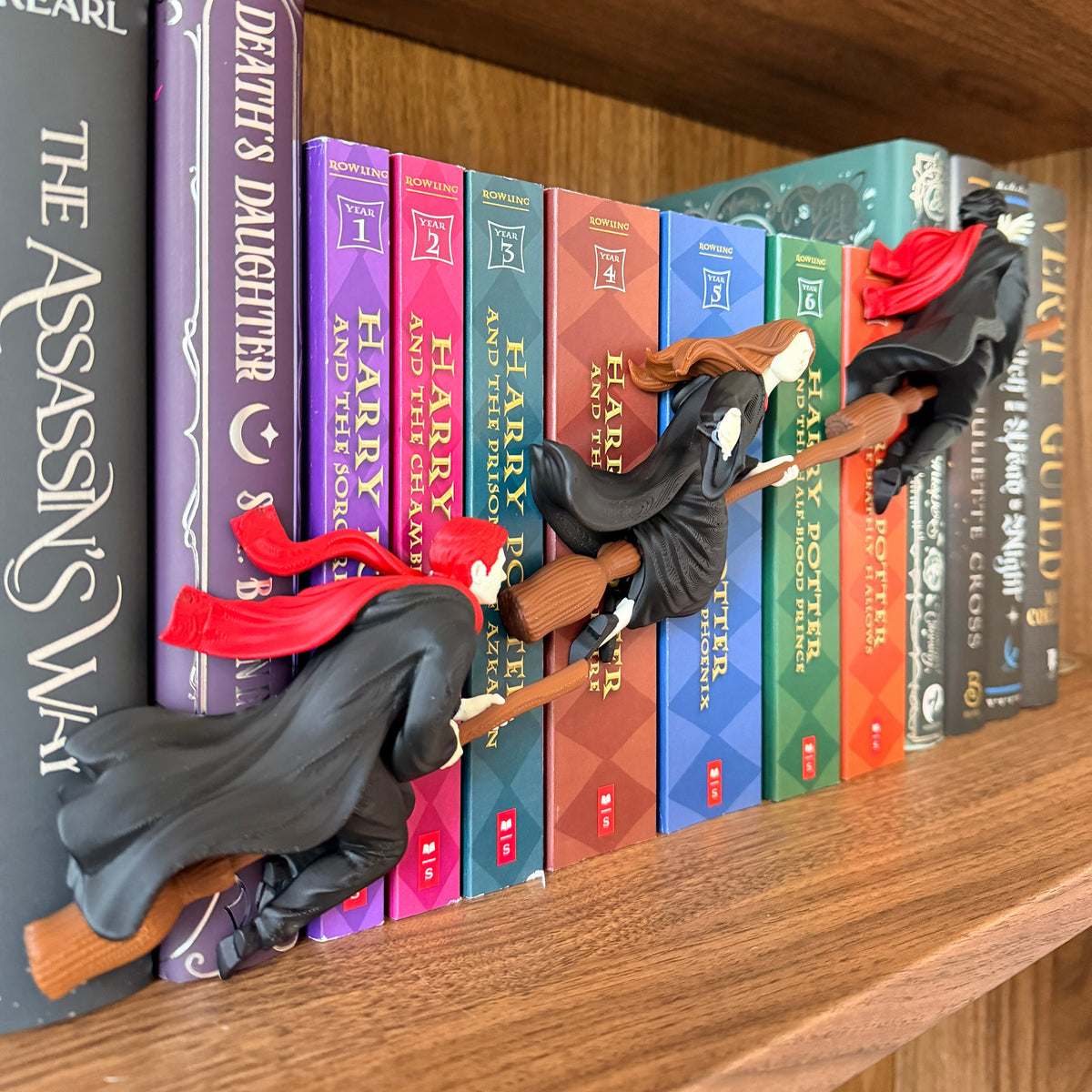 A row of Harry Potter book spines arranged on a wooden shelf, with small witch figurines in black robes and red hats perched on the books.
