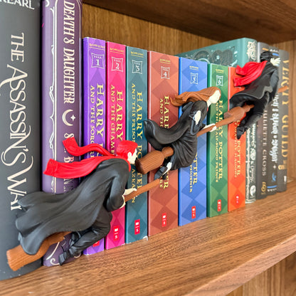 A row of Harry Potter book spines arranged on a wooden shelf, with small witch figurines in black robes and red hats perched on the books.