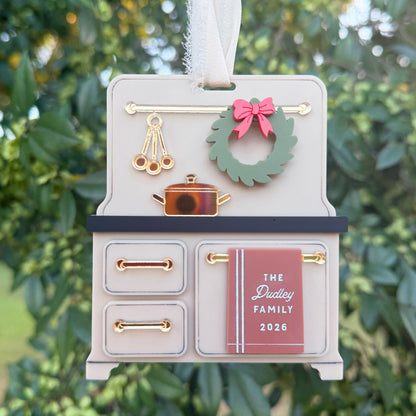 A miniature kitchen-themed Christmas ornament shaped like a small stove, held by a hand against a blurred outdoor background of green foliage. The ornament is light gray with gold handles and knobs, a green wreath with a pink bow, and a pink door labeled 'The Duttley Family 2026'.