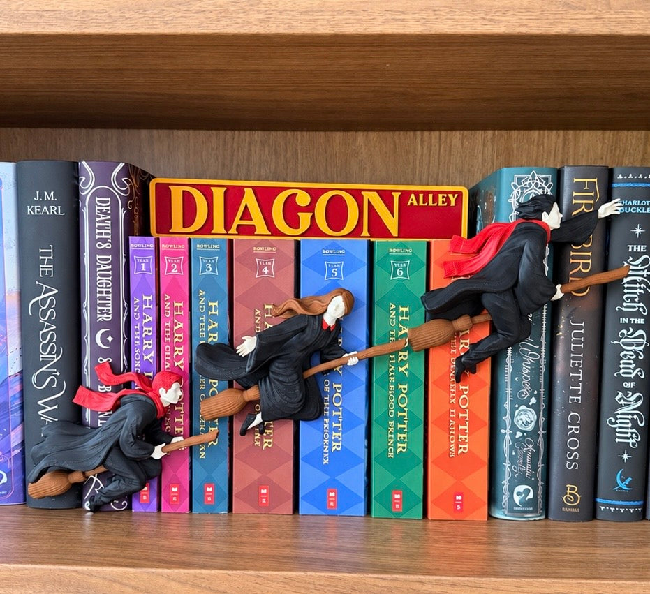 Three small witch figurines in black robes with red scarves are arranged on a wooden bookshelf between colorful Harry Potter books, including Diagon Alley and Harry Potter titles.