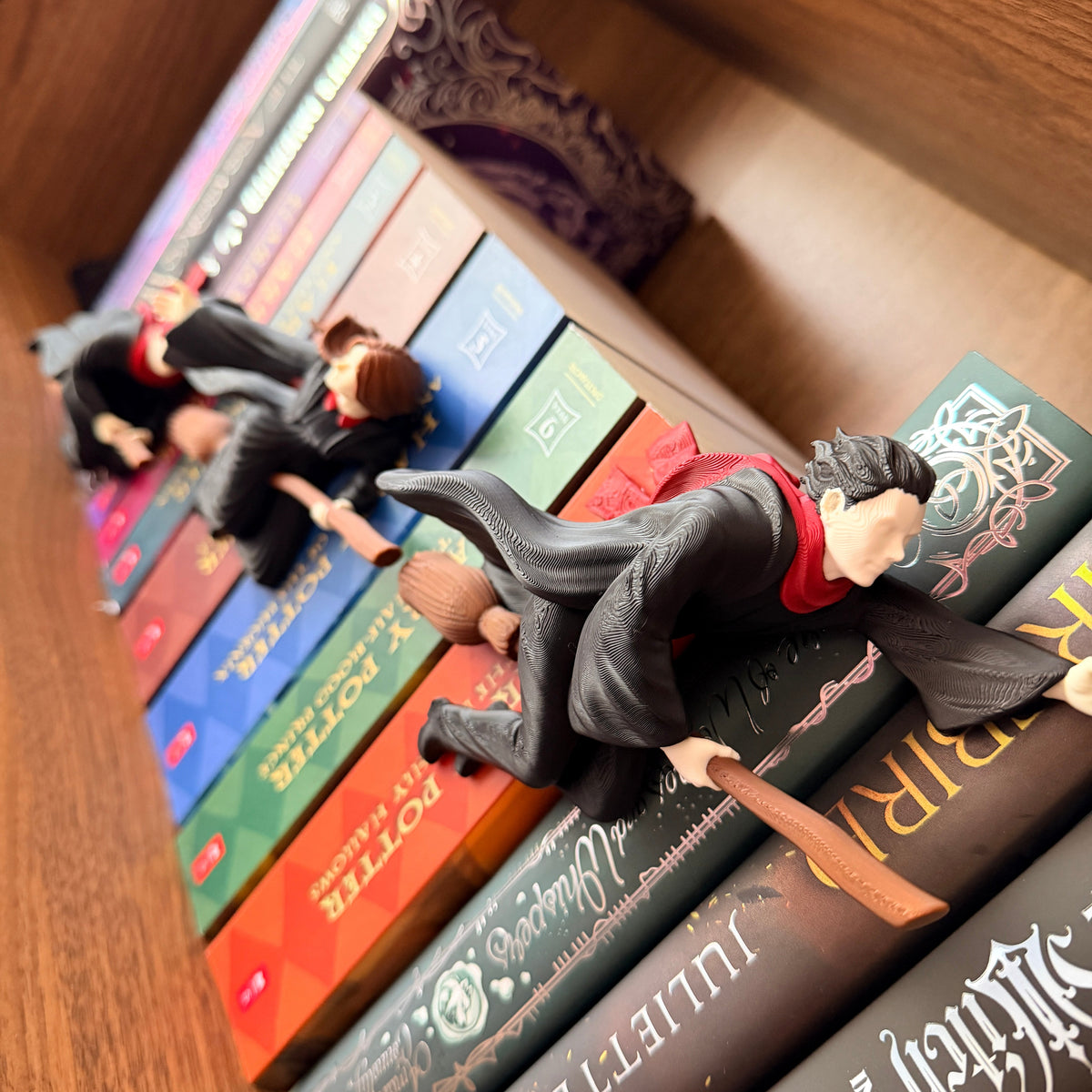 Three small figurines of Harry Potter characters lying on top of a row of colorful Harry Potter books on a wooden shelf.