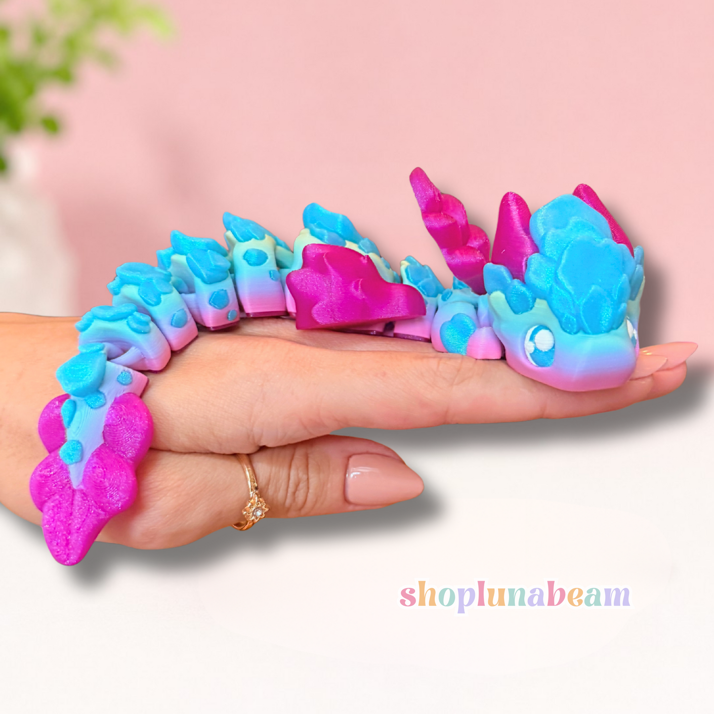 A colorful dragon toy draped over a person's hand, featuring a gradient of blue, pink, and purple, set against a soft pink background.