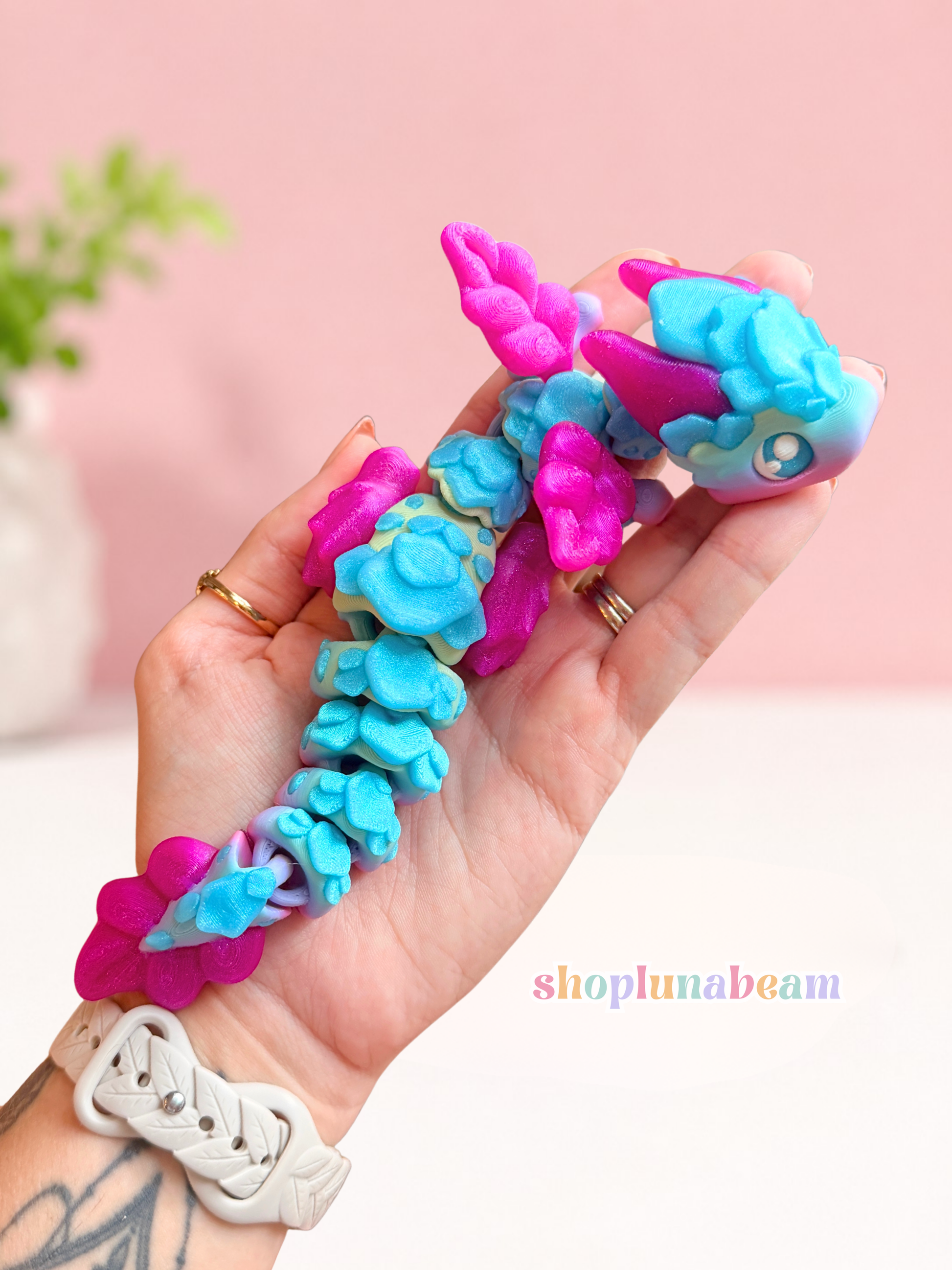 A hand holding a small articulated toy dragon in bright turquoise blue and magenta pink, with a soft pink background and a visible shoplunabeam watermark.