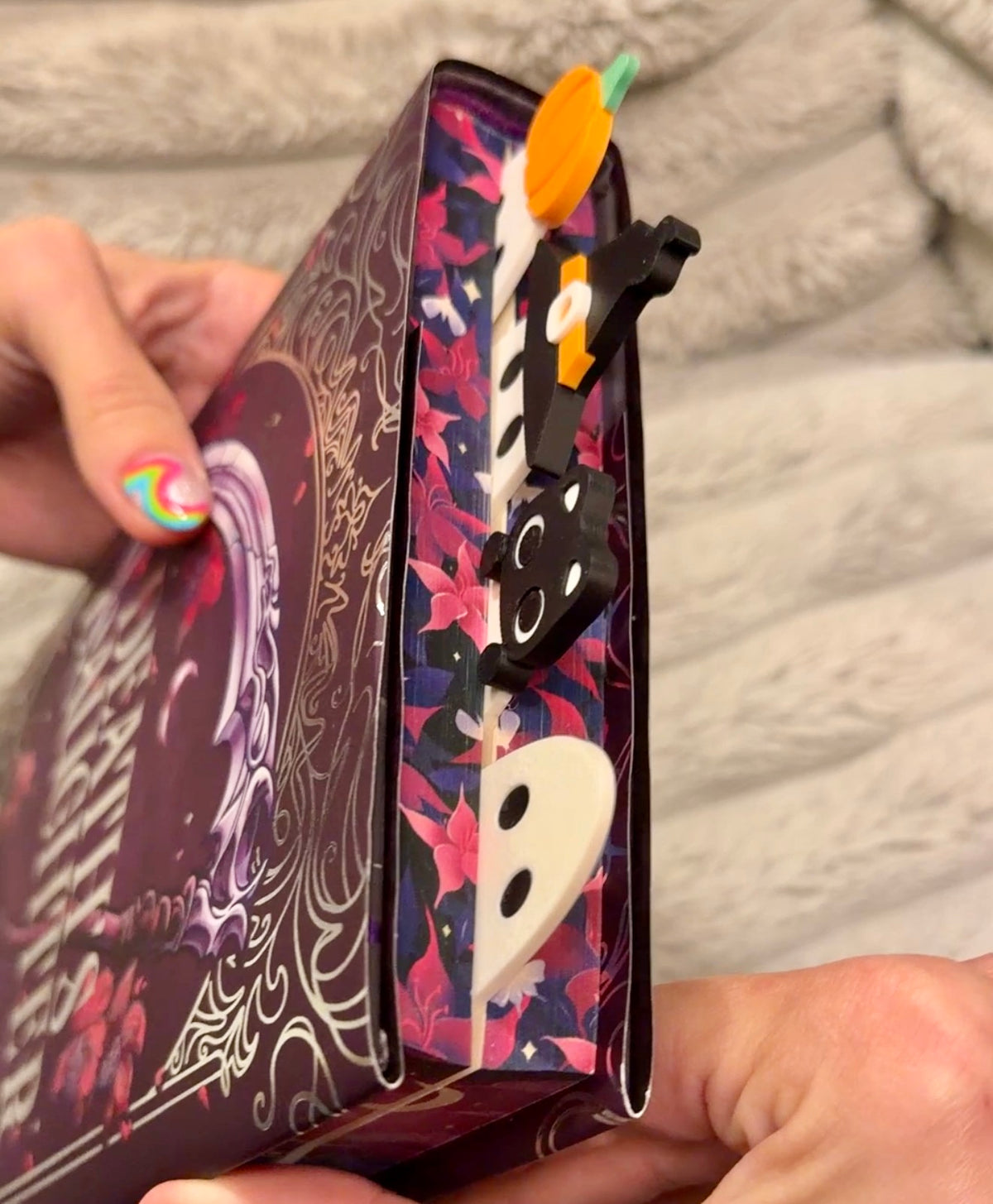 A close-up of a person holding a decorative book or notebook with a Halloween-themed cover. The cover features a dark purple and black base with floral patterns, white gothic swirls, and cartoonish ghost and pumpkin bookmarks sticking out.
