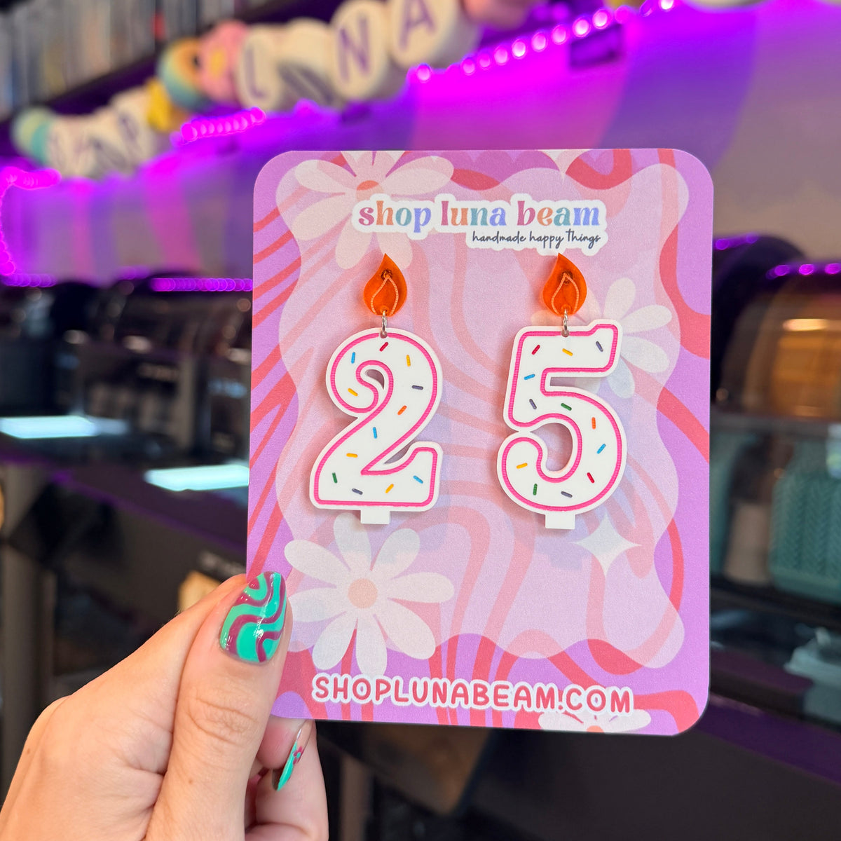 A hand with colorful nail art holds a pink card displaying two novelty candle-shaped earrings shaped like the numbers 2 and 5, with flame tops. The card reads 'shop luna beam' and 'SHOPUNABEAM.