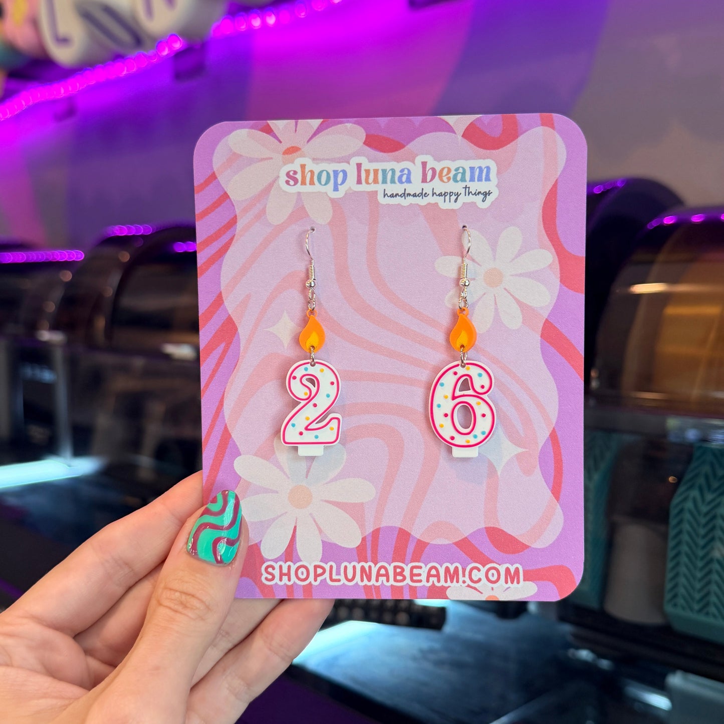 A hand holds a pink display card featuring a pair of dangling earrings shaped like the numbers 2 and 6, with orange beads and silver hooks. The card is branded 'shop luna beam'.
