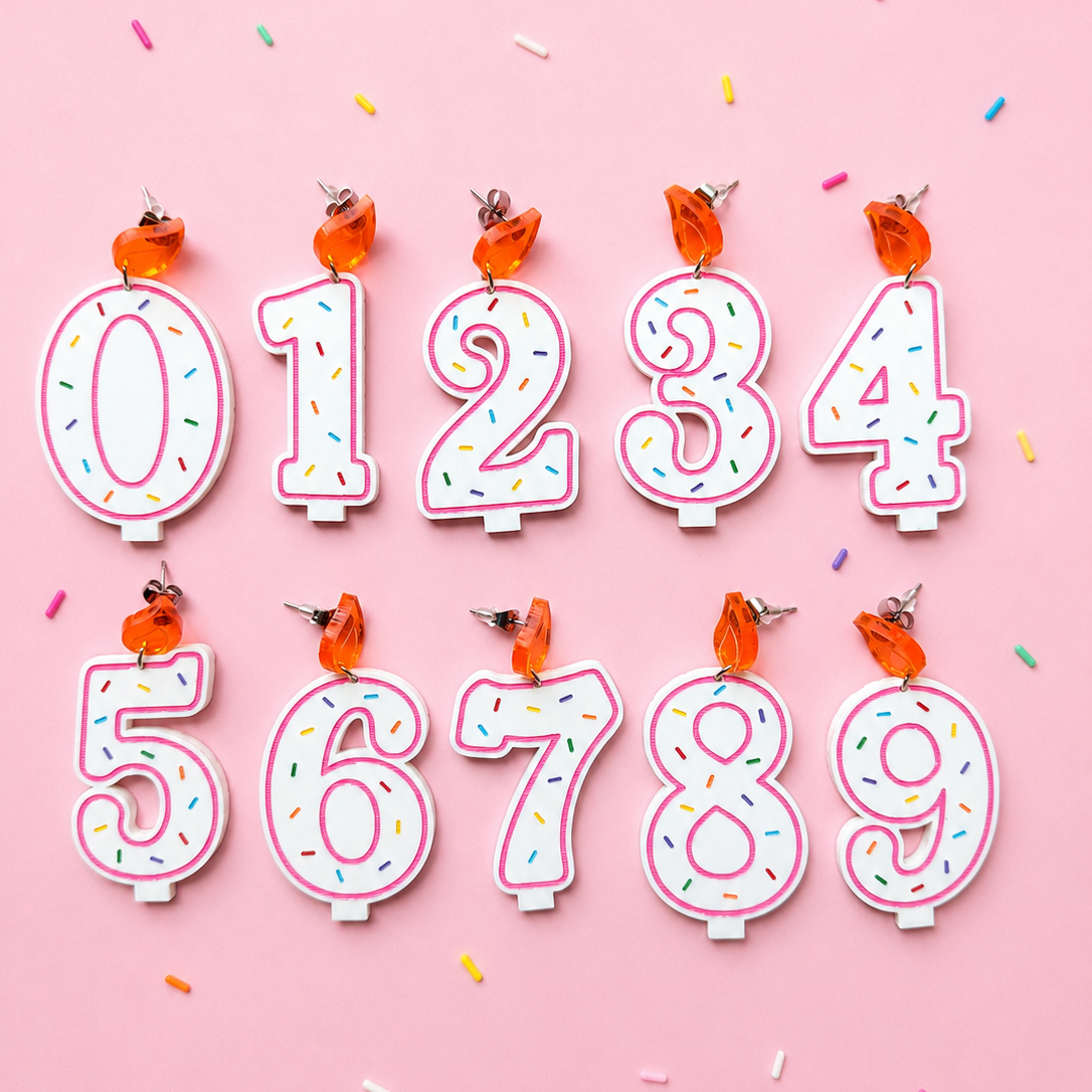 A set of nine birthday candles shaped like the numbers 0 through 9, decorated with white frosting-like tops and colorful sprinkles, arranged on a pink background with small confetti pieces.
