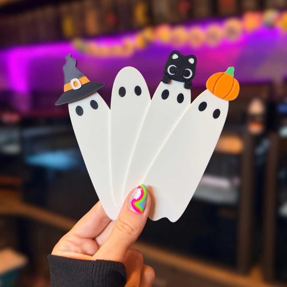 A hand holds four white ghost-shaped bookmarks or cards with Halloween-themed tops: a gray witch hat, a black cat, and an orange pumpkin, against a blurred indoor background with purple lighting.