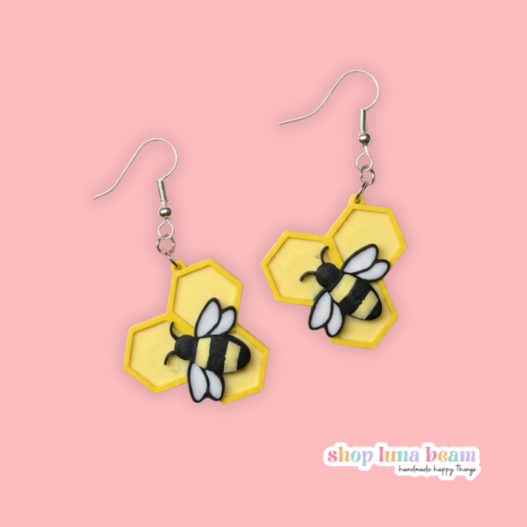 A pair of handmade dangle earrings featuring yellow honeycomb shapes with black and white bumblebees, suspended from silver fishhook wires, displayed on a solid pink background with a 'shop luma bean' logo in the bottom right corner.