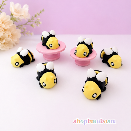 Five small bumblebee-shaped accessories arranged on a pale pink surface against a lavender background, with two displayed on pink stands. Decorative flowers appear in the upper left, and a colorful 'shoplunabeam' watermark is at the bottom right.