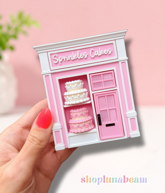 A miniature pink and white bakery shop figurine labeled 'Sprinkles Cakes' held in a hand with red nail polish, set against a soft pink background.