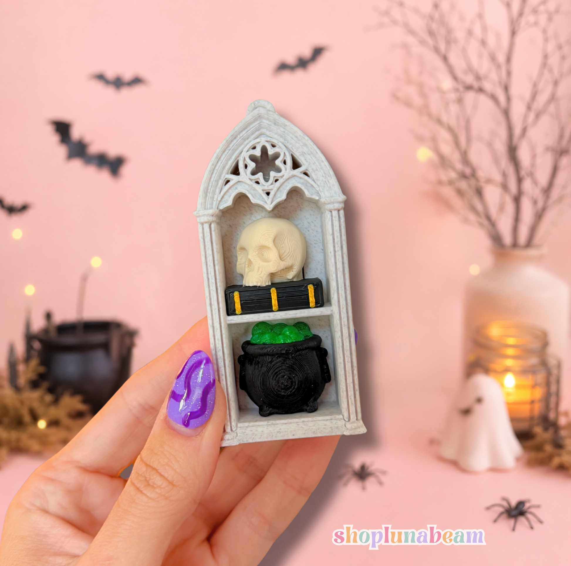 A hand with purple nail art holds a miniature gothic shelf ornament featuring a skull and a black cauldron with a green gem, set against a pink Halloween-themed background with bats and a ghost.