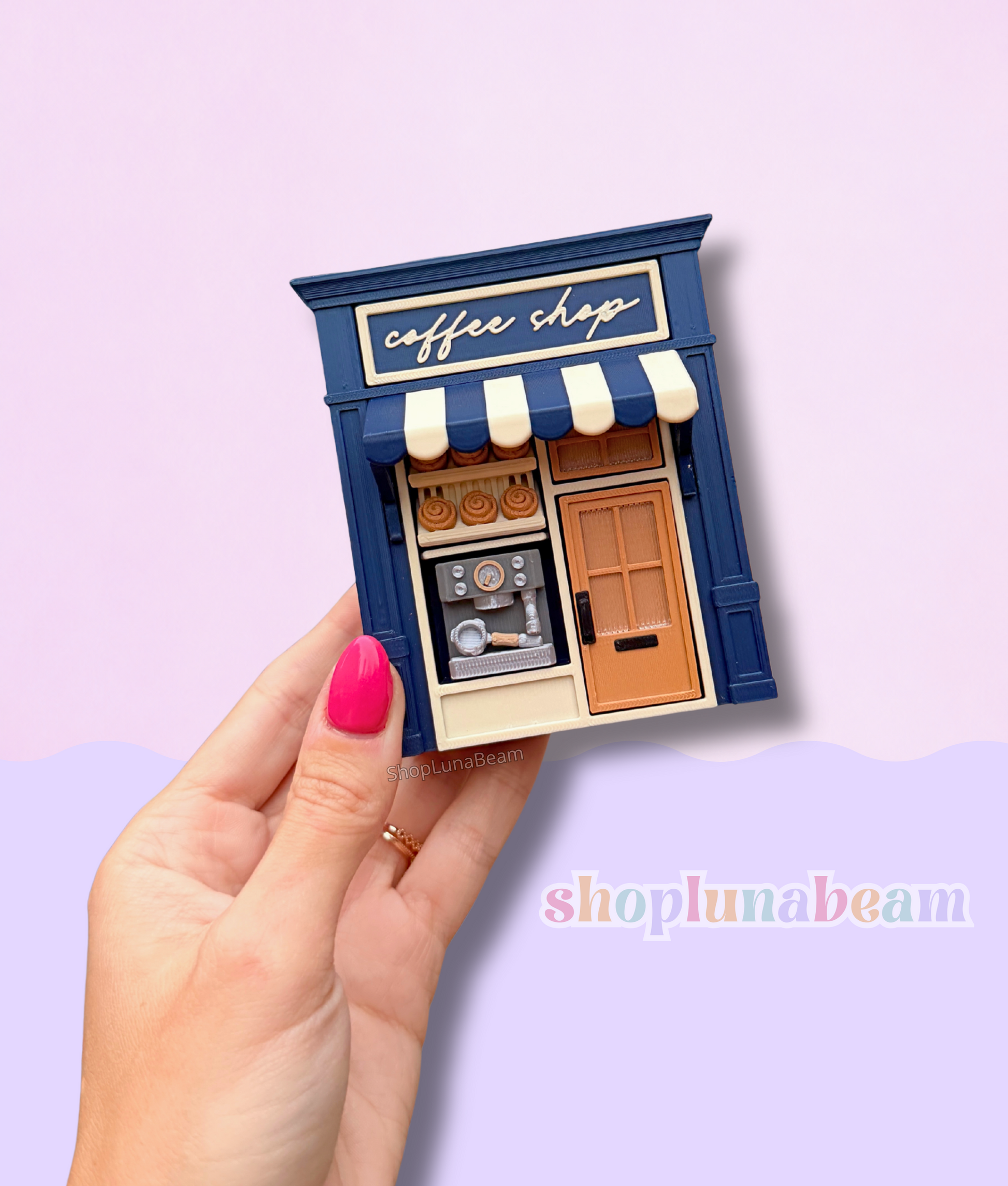 Miniature model of a coffee shop with a hand holding it against a light purple background.