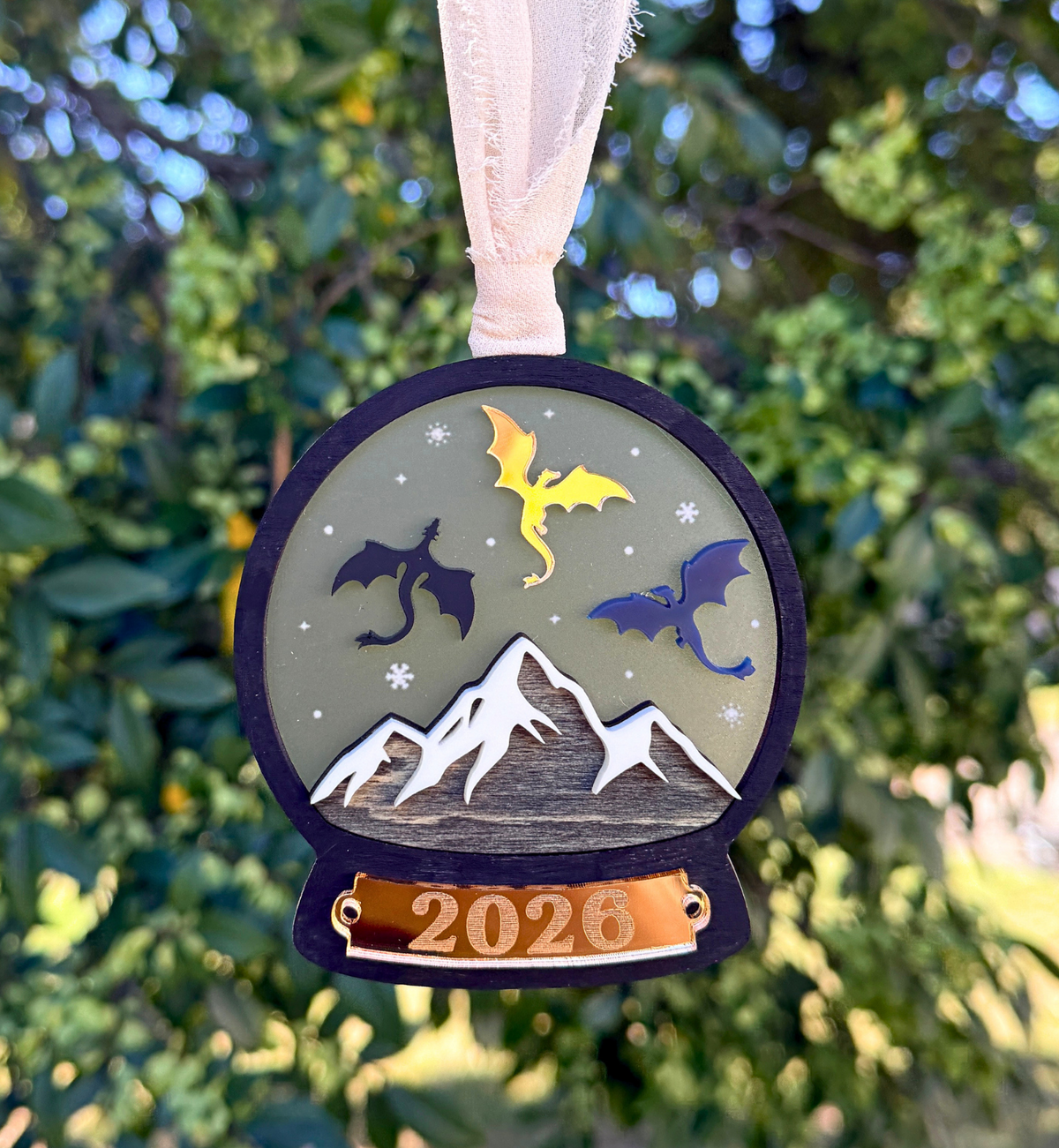 A round hanging ornament with a white ribbon, featuring three stylized dragons above white mountains and the year 2026 on a gold banner, photographed outdoors against a blurred green tree background.