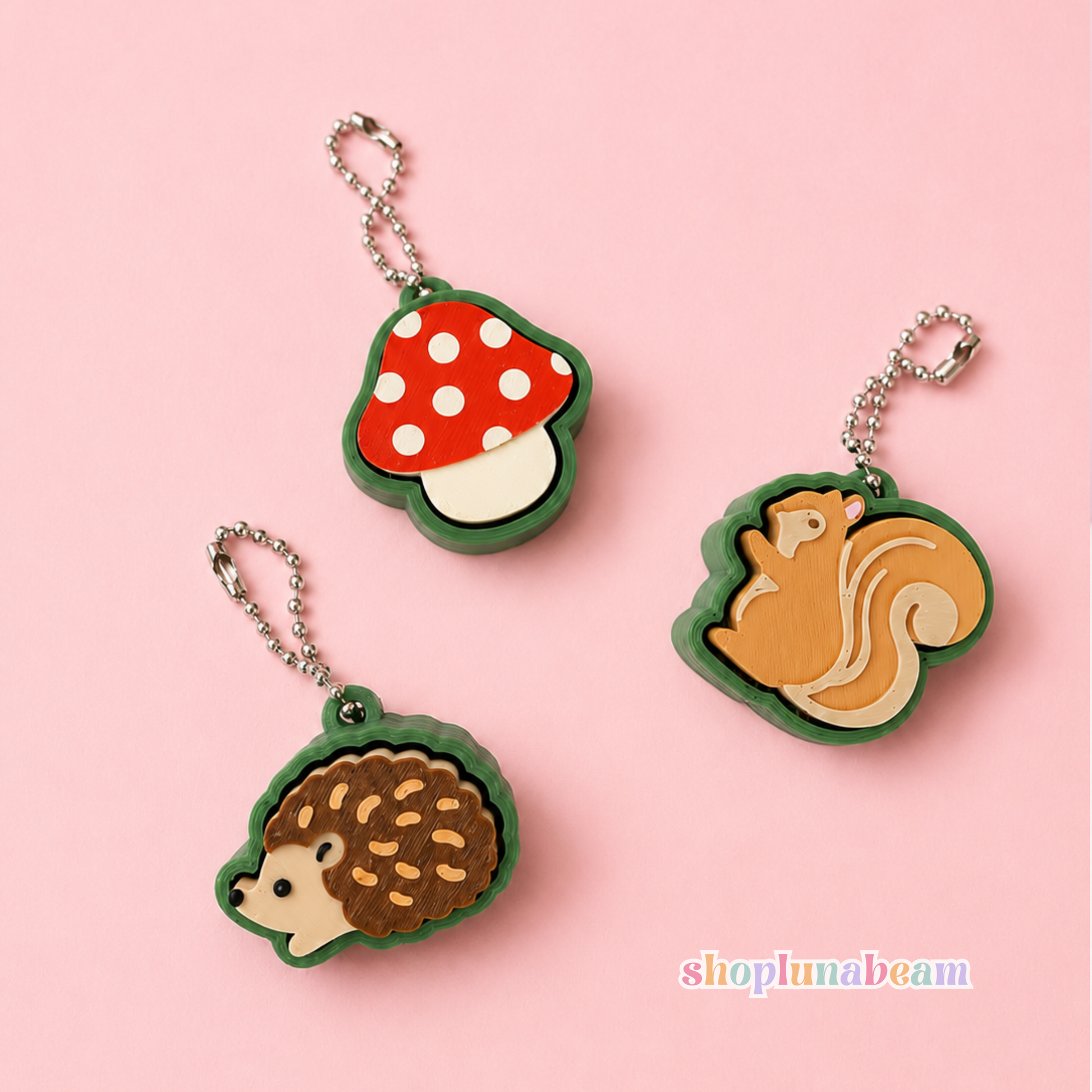 Three small animal and nature-themed keychains or charms on a light pink background: a red-and-white mushroom with green border, a brown hedgehog with green border, and a tan-and-cream squirrel with green border. Each charm has a silver chain loop.