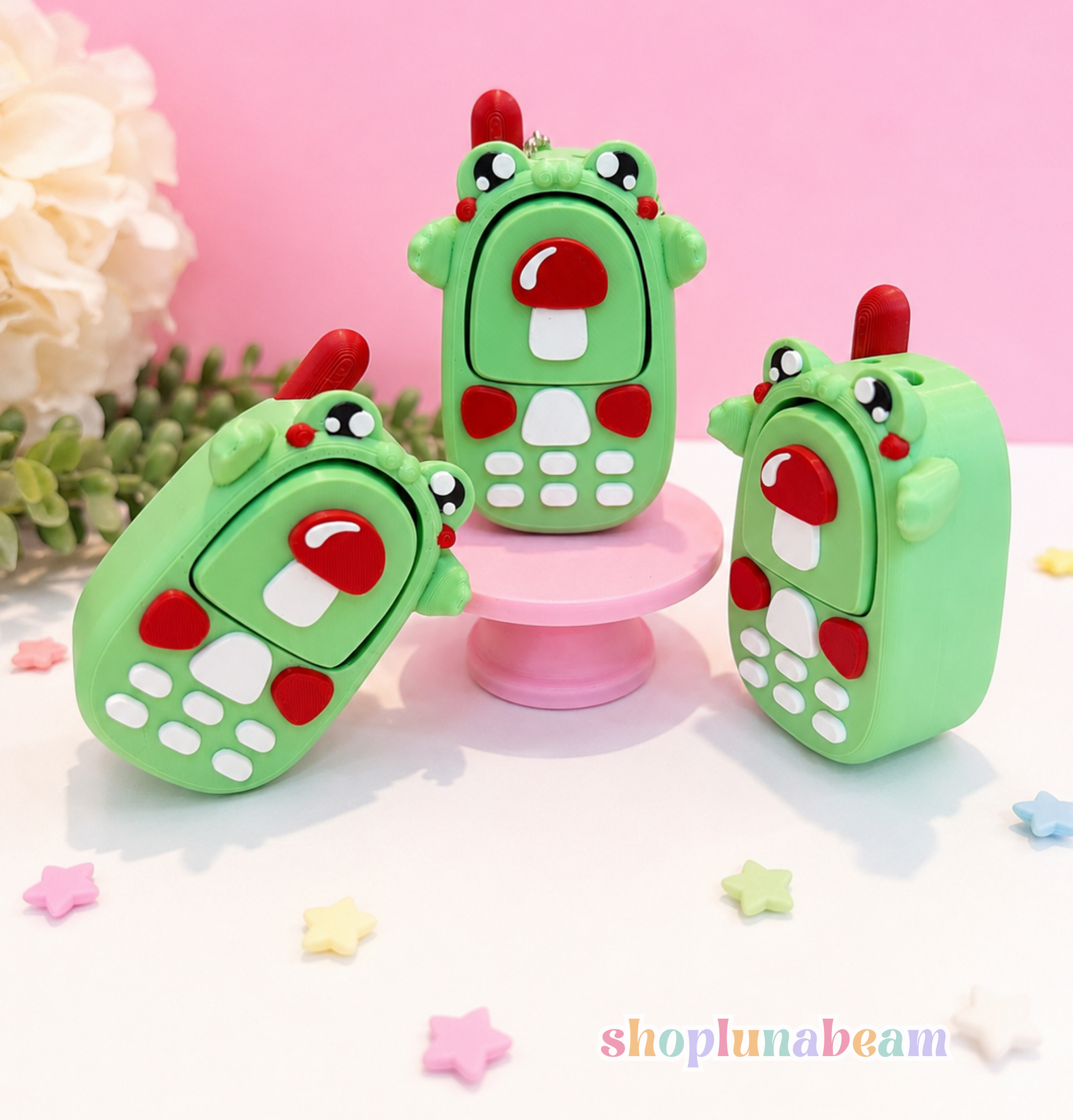 Three green frog-themed USB flash drives styled like mobile phones, arranged on a white surface with a pink background and decorative flowers.