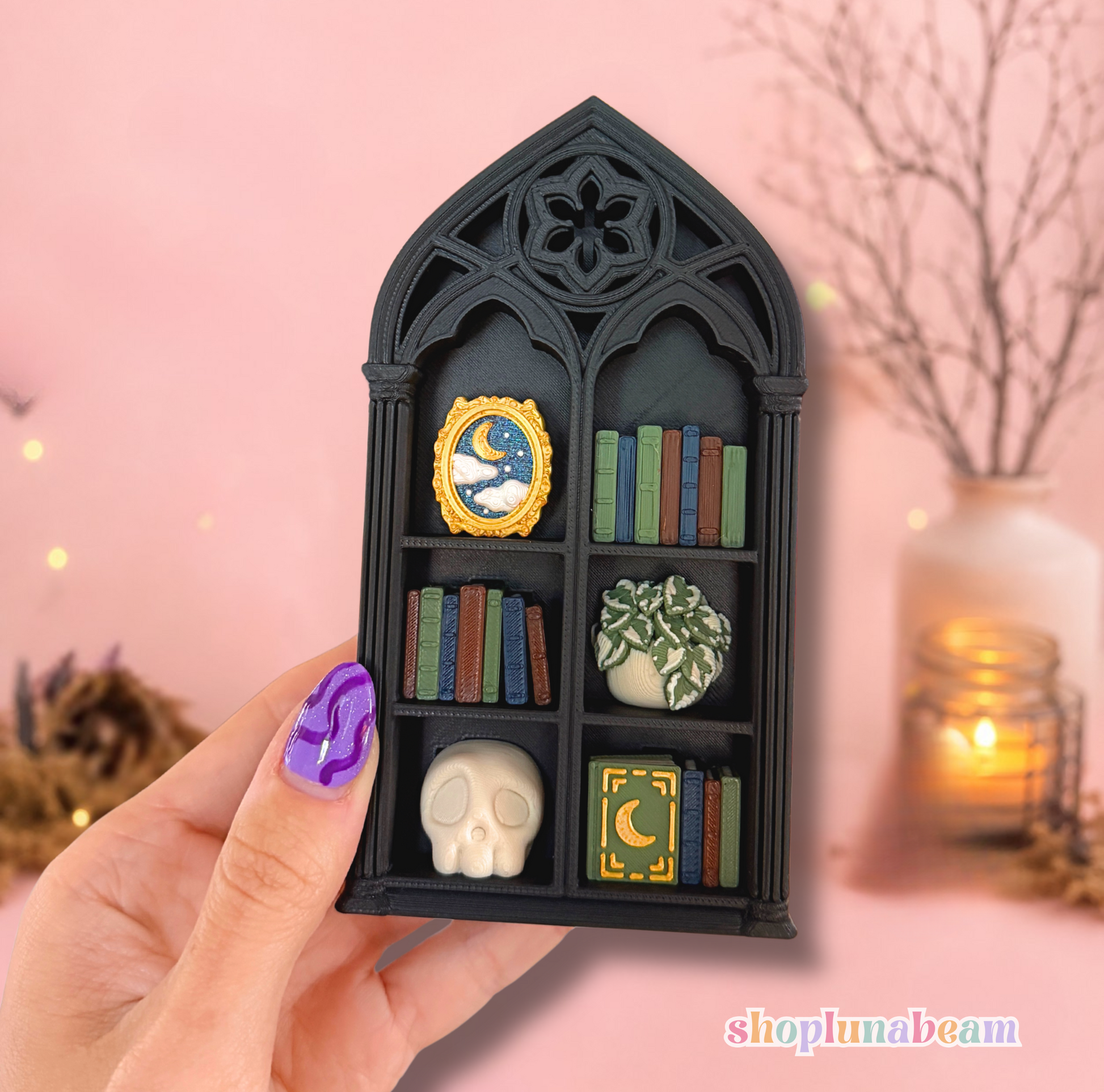 A hand with purple nail art holds a miniature black gothic arched shelf filled with tiny decorative items, including books, a skull, a small plant, and a moon-themed ornament, against a soft pink background with blurred holiday lights and a vase.