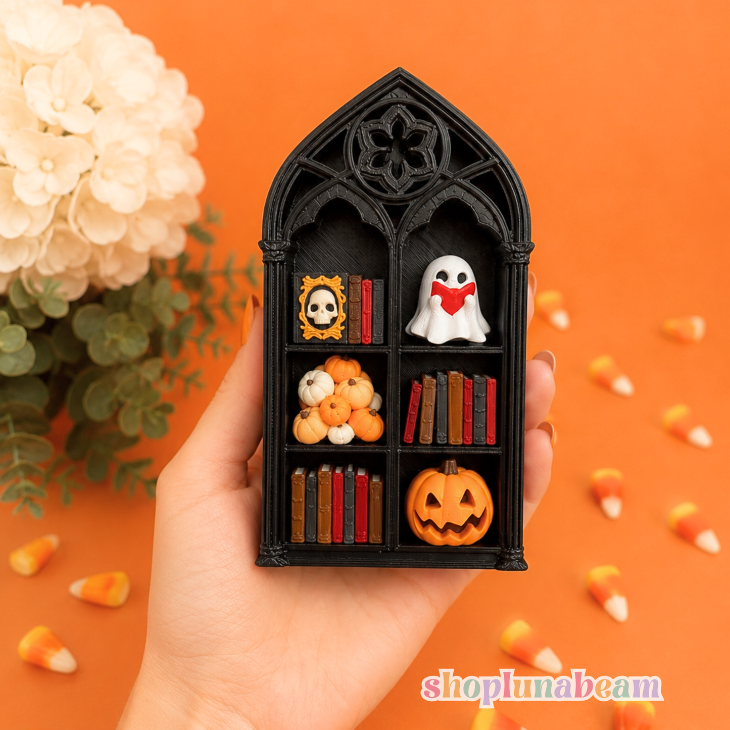 A hand holds a miniature black gothic window bookshelf ornament decorated with tiny Halloween figurines, including a ghost, pumpkin, books, and a skull, against an orange background with scattered candy corn and a white flower.