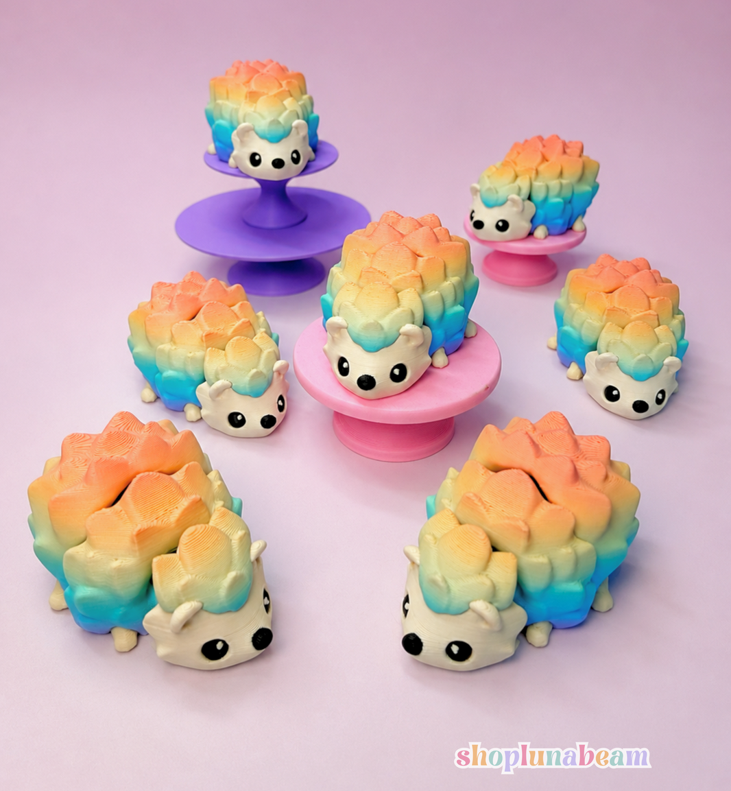 Six small pastel hedgehog-shaped items arranged on a pink and lavender studio background, with three displayed on standees. The items have a rainbow gradient from peach/orange to blue and yellow, with white faces and black eyes.