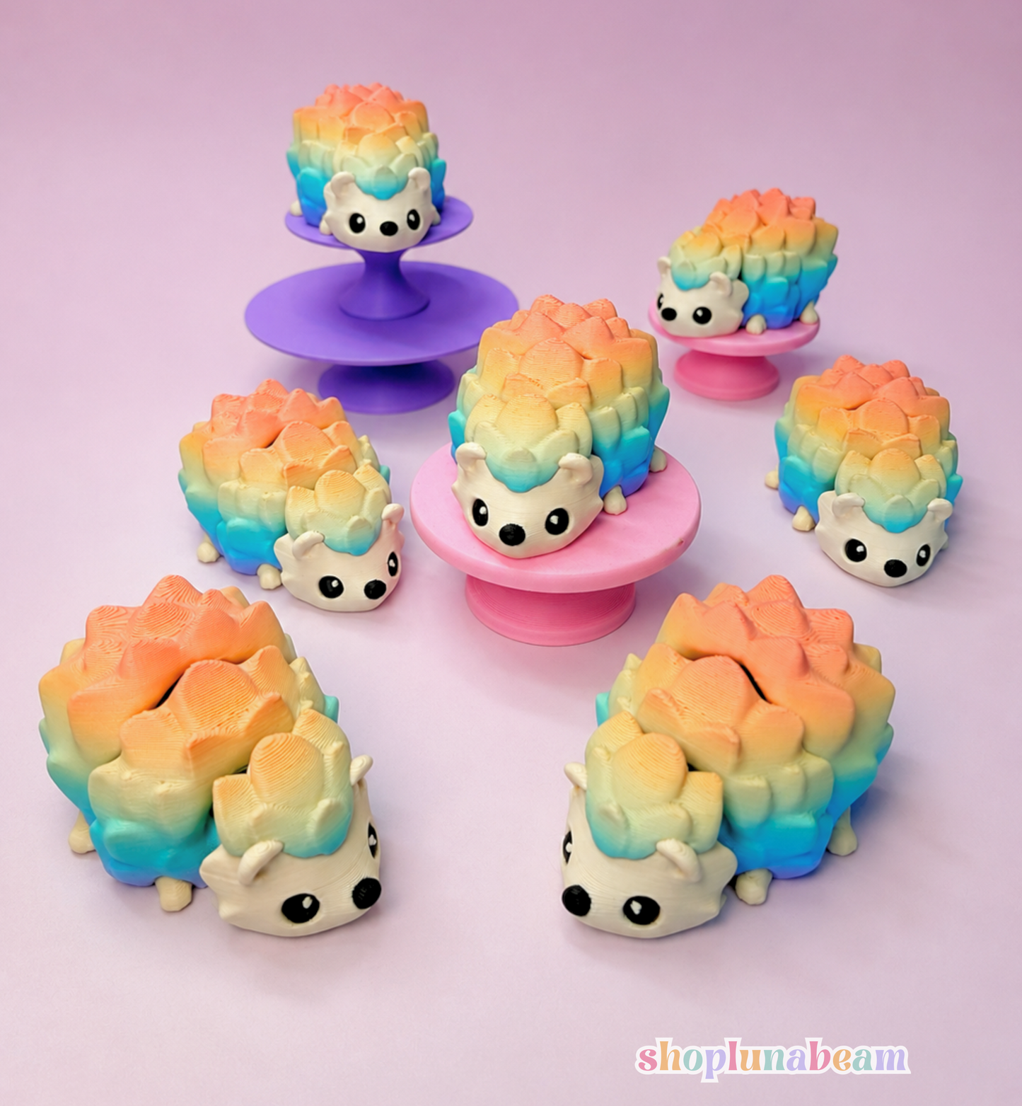 Six small pastel hedgehog-shaped items arranged on a pink and lavender studio background, with three displayed on standees. The items have a rainbow gradient from peach/orange to blue and yellow, with white faces and black eyes.