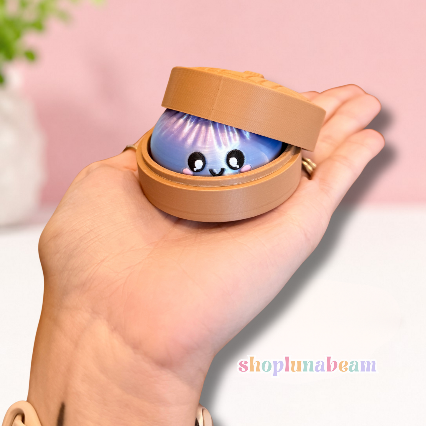 A hand holding a small round tan container with a lid that is slightly open, revealing a cute blue and purple character face peeking out. The background is a soft pink wall with a blurred green plant in the corner.