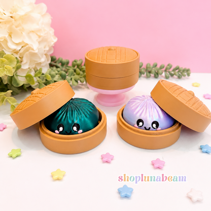 Three cute bun-shaped cosmetic containers in peach-brown, shown open to reveal colorful, character-like contents: one teal with black eyes, one lavender with a smiley face, and one closed in the background. Decorative flowers, greenery, and pastel star confetti are arranged on a pink and white surface.