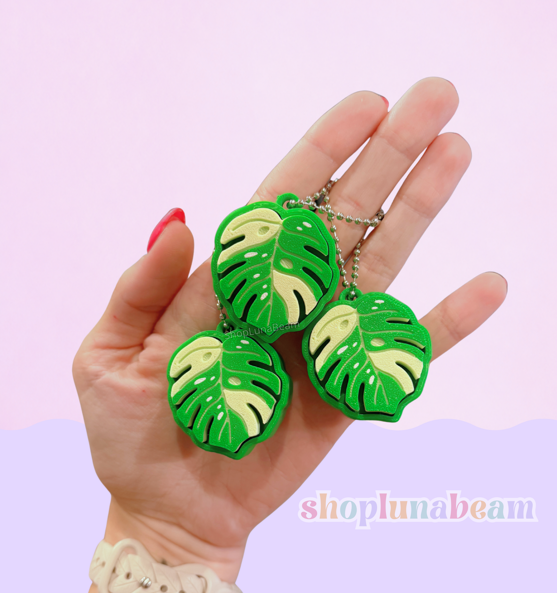 Hand holding three green leaf-shaped keychains against a light pink background with 'shopplumabeam' branding.