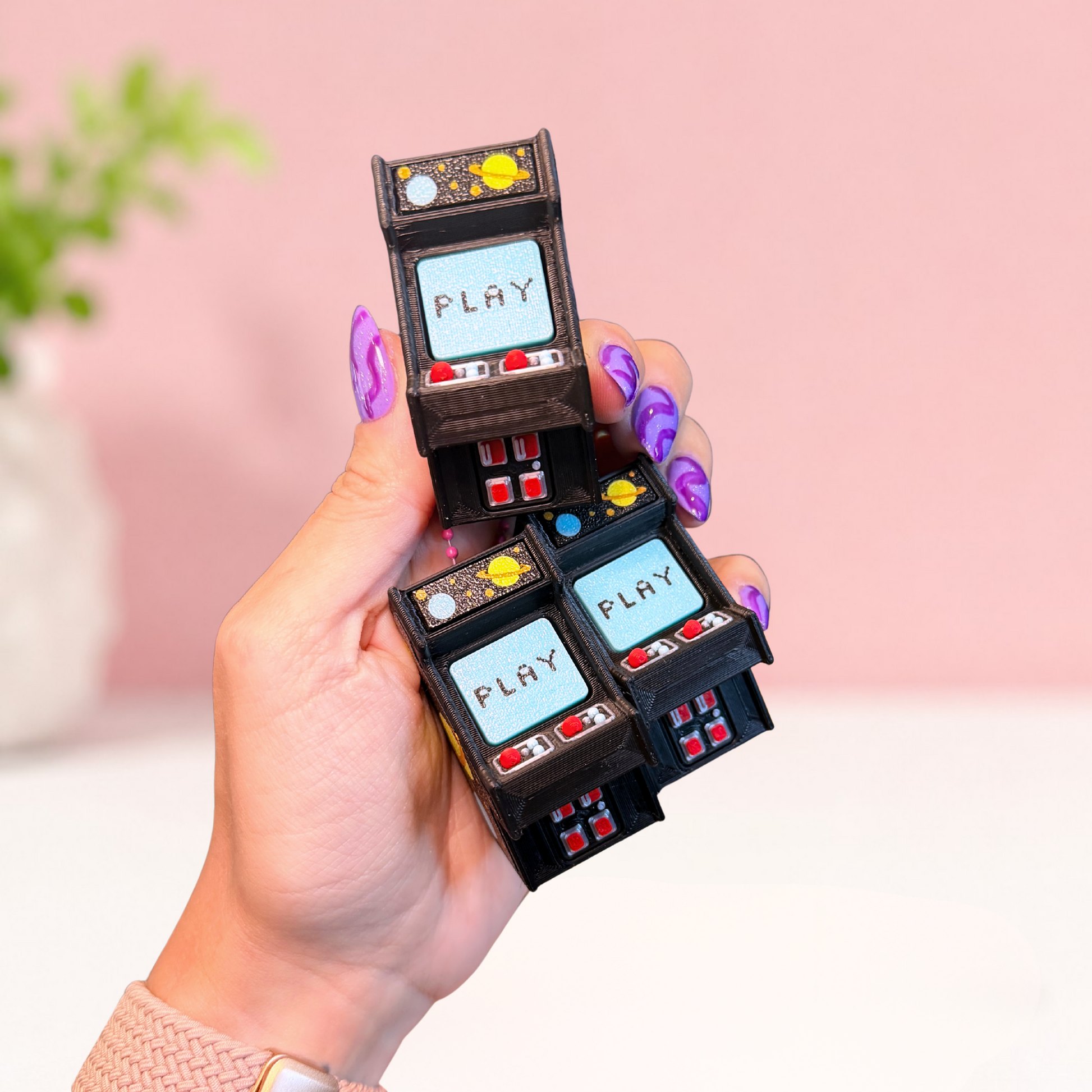 A hand with purple nail art holds three miniature black arcade machine charms with light blue screens displaying the word 'PLAY'. The background is a soft pink and white studio setting with a blurred plant.