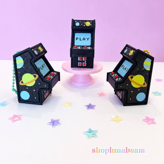 Three miniature black arcade machine keychains decorated with colorful space-themed graphics, including planets and stars, displayed on a white surface with pastel star stickers and a pink background.