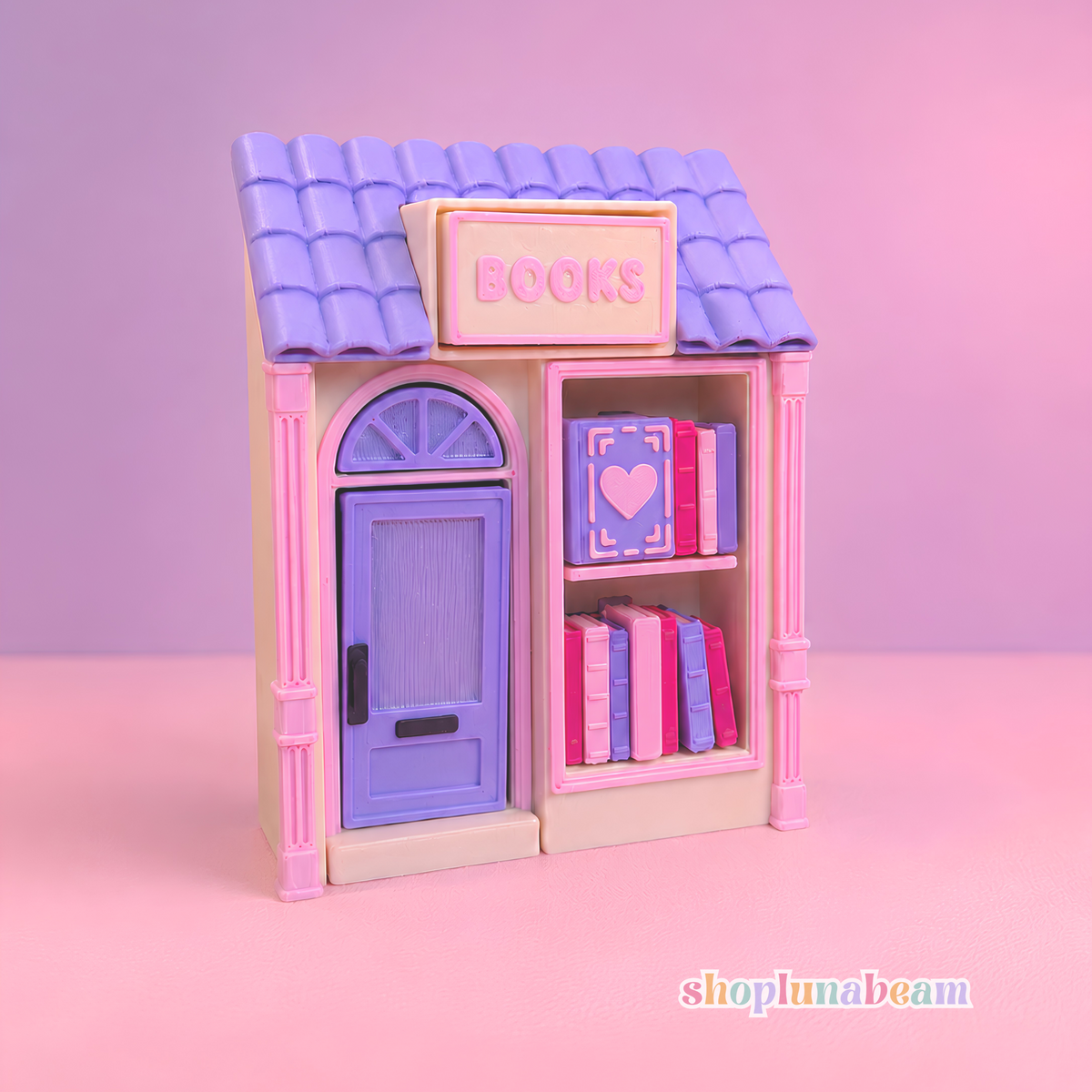 A miniature dollhouse-style bookshop with a lavender purple tiled roof, pink trim, a lavender door, and shelves filled with colorful books. The word 'BOOKS' is printed on the front above the door.