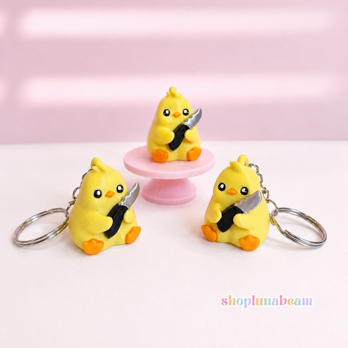 Three small yellow duck keychains with orange feet and black-and-white striped beaks, each holding a silver knife, displayed on a white surface with a pink background. One keychain is placed on a pink pedestal.