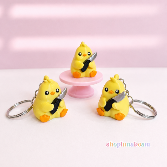 Three small yellow duck keychains with orange feet and black-and-white striped beaks, each holding a silver knife, displayed on a white surface with a pink background. One keychain is placed on a pink pedestal.