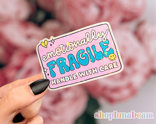 Sticker with 'Emotionally Fragile Handle with Care' text held by a hand, with a blurred background.