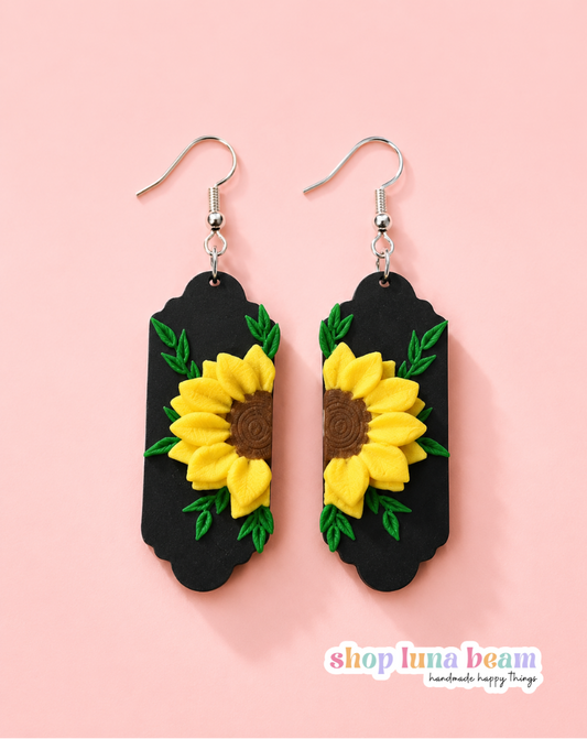 A pair of handmade dangle earrings featuring yellow sunflowers with brown centers and green leaves on a black background, suspended from silver fishhook wires, set against a solid pink background.