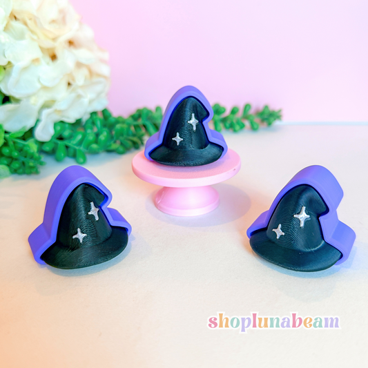 Three small wizard hat-shaped accessories in purple and black with white star details, displayed on a light surface with a pink background and decorative flowers.