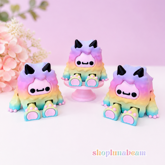 Three cute pastel rainbow monster figurines with black cat ears and simple face details, displayed on a white surface against a soft pink background with decorative flowers.